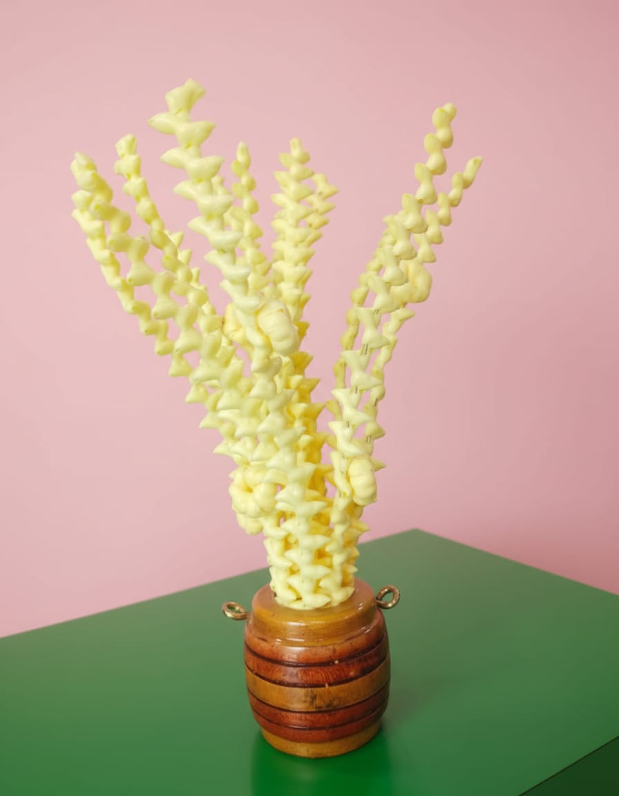 A small, barrel-shaped handcrafted wooden planter filled with textured, bright yellow decorative stems, sitting on a green surface against a pastel pink background.