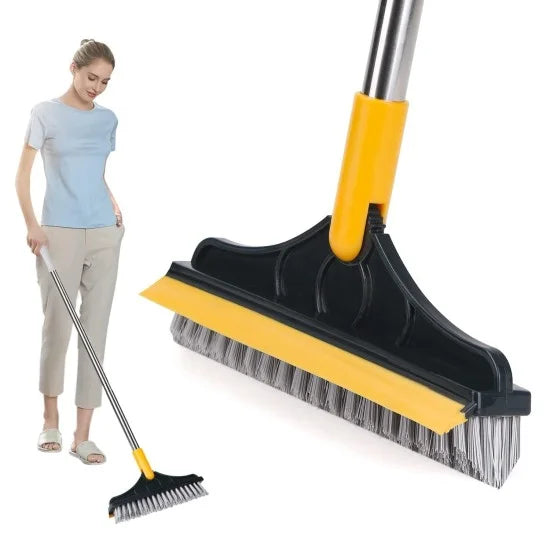 A close-up of a 2-in-1 floor cleaning brush head featuring stiff grey bristles and a yellow rubber squeegee edge, with a woman using the long-handled tool in the background.