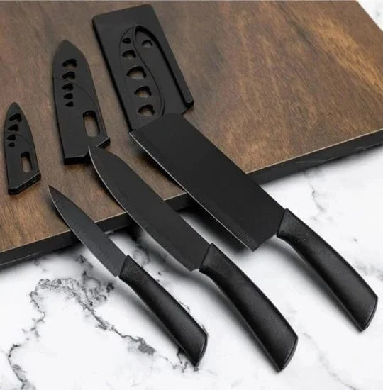 A set of three matte black kitchen knives of varying sizes, including a paring knife, utility knife, and cleaver, displayed on a white marble surface alongside their matching black protective sheaths resting on a wooden cutting board.