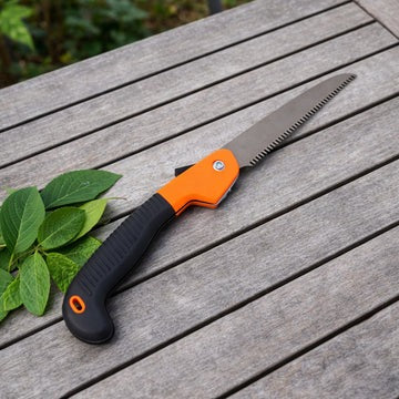A folding pruning saw in the open position, featuring a silver toothed blade and a textured black and bright orange handle, resting on a slatted wooden table beside a small sprig of green leaves.