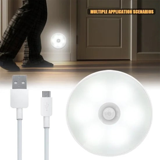 A close-up of a white, circular motion-sensor LED night light and its USB charging cable. In the background, the same light is shown mounted low on a hallway wall, illuminating the floor as a person wearing pajamas walks past in the dark.