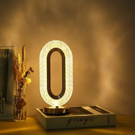 An elegant oval-shaped crystal LED table lamp emitting a warm ambient glow, resting on a decorative book next to dried flowers.