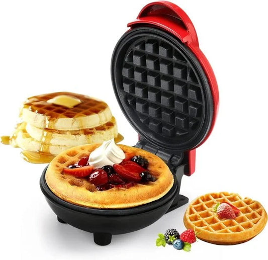 An open red mini waffle maker displaying a freshly cooked round waffle topped with berries and cream, with a stack of syrup-drizzled waffles in the background.