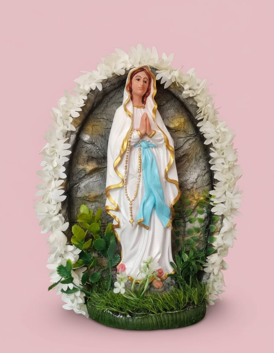 A statue of Mother Mary with hands folded in prayer, wearing a white robe and blue sash, standing inside a stone-like grotto framed by white flowers and green foliage.