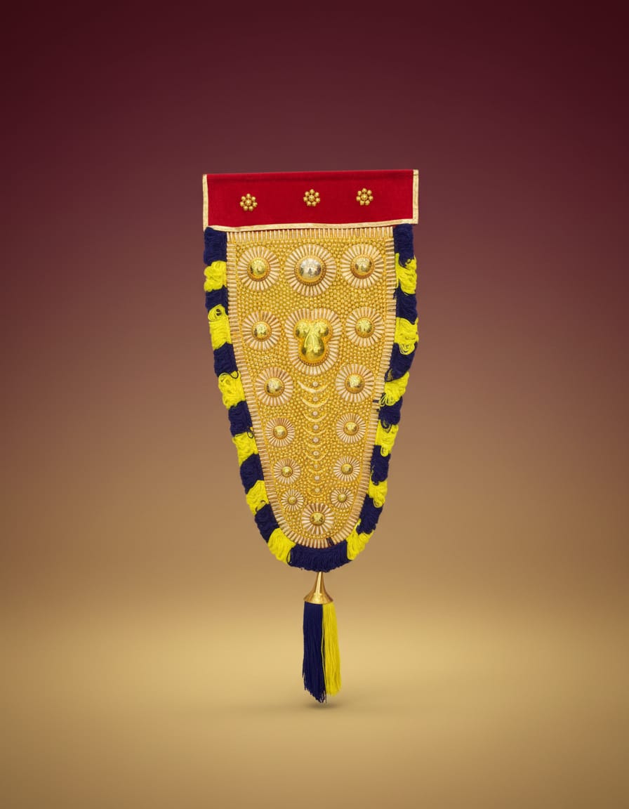 A traditional Kerala Nettipattam wall hanging featuring an intricate golden centerpiece bordered by a red band at the top and black and yellow fringed edges, finishing with a bottom tassel.