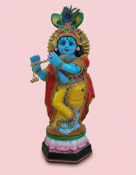 A colorful handpainted idol of Lord Krishna playing a golden flute, featuring a bright blue body, yellow and red garments, and a peacock feather crown, standing on a pink and green lotus base.