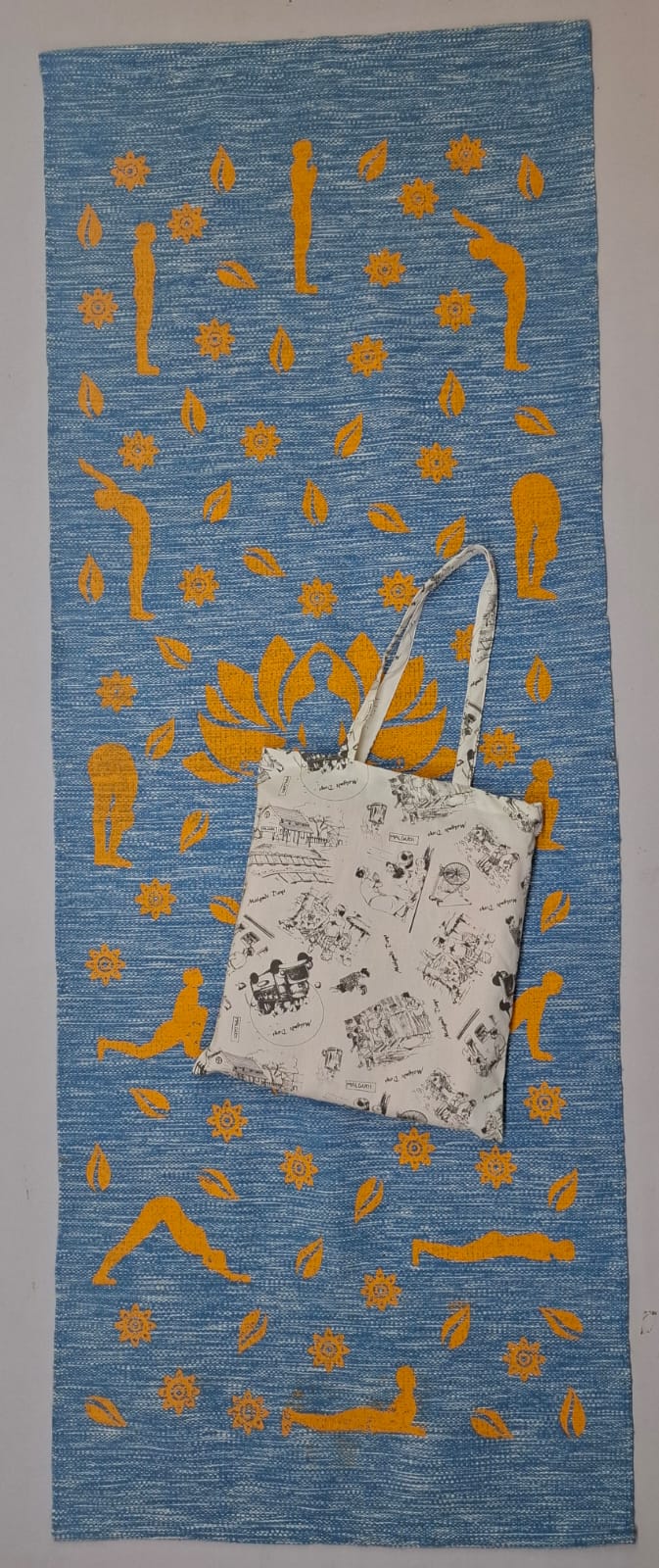 A blue eco-friendly cotton yoga mat printed with yellow Surya Namaskar poses, shown with a matching printed light-colored tote bag resting on top.