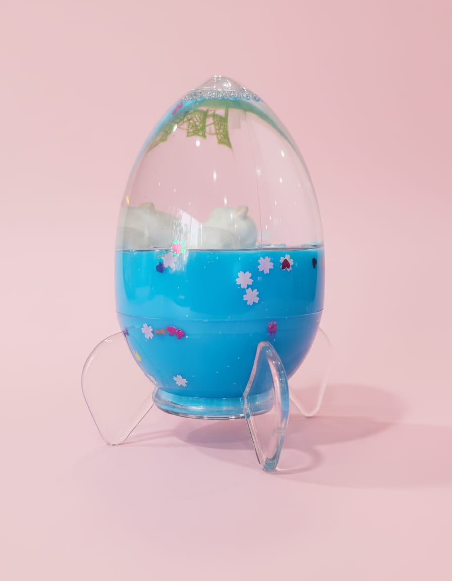 A cute rocket-shaped snowglobe with a blue base and clear fins, featuring floating star confetti inside, displayed against a soft pink background.