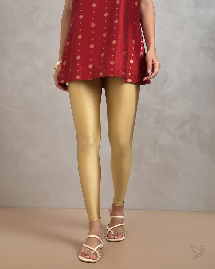 A woman modeling a pair of shiny gold ankle-length leggings paired with a red patterned top and white strappy sandals.